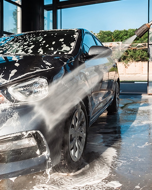 Lavage voiture haute pression : mousse et eau sur carrosserie Voiture foncée lavée avec un nettoyeur haute pression, de la mousse blanche recouvrant le capot et le pare-brise, sol mouillé.