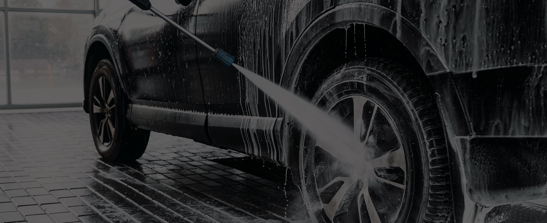 Lavage voiture haute pression : mousse et eau sur carrosserie Voiture foncée lavée avec un nettoyeur haute pression, de la mousse blanche recouvrant le capot et le pare-brise, sol mouillé.