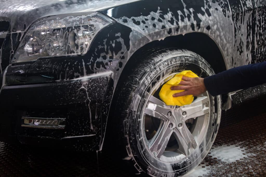Lavage de Jante et Pneu : Nettoyage de Voiture Moussé Lavage de jante et pneu Continental d'un véhicule noir couvert de mousse, une main essuie avec un chiffon jaune.