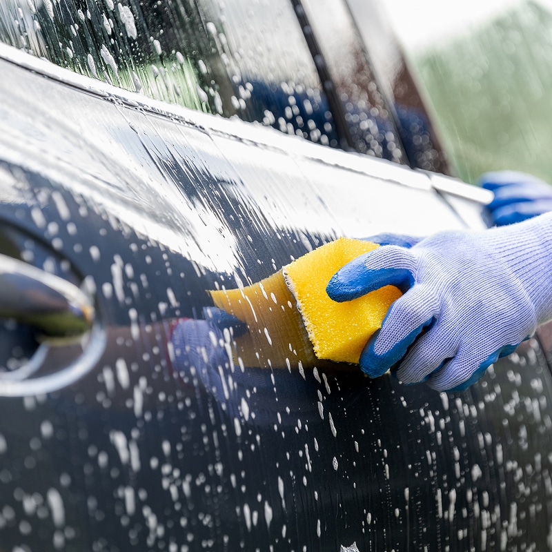 Lavage voiture : Main avec gant nettoyant l'extérieur Gant bleu lavant une carrosserie sombre et mouillée avec une éponge jaune et de la mousse.