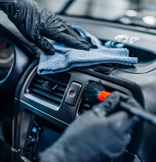Nettoyage intérieur voiture : détails et finition pro Gros plan mains gantées nettoyant tableau de bord voiture avec chiffon bleu et brosse douce autour d'une grille d'aération et d'un bouton d'urgence.