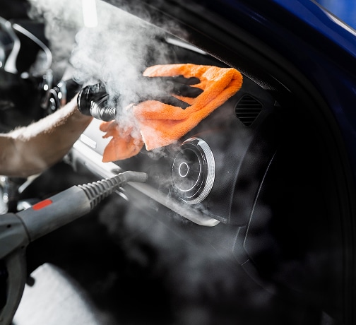 Désinfection Vapeur Intérieur Voiture : Nettoyage Profond Nettoyage vapeur intérieur voiture : un ganté essuie le tableau de bord près d'une aération chromée avec une serviette orange et un pistolet vapeur.