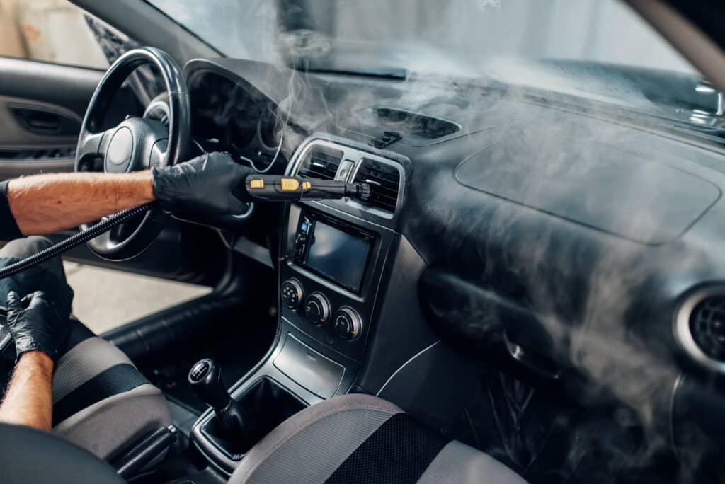 Nettoyage vapeur intérieur voiture : désinfection et detailing Homme ganté utilisant un nettoyeur vapeur sur le tableau de bord d'une voiture, dégageant de la vapeur autour du volant et de la console centrale.