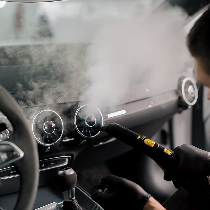 Nettoyage vapeur intérieur voiture: désinfection des aérateurs Détail gros plan sur l'intérieur d'une voiture Audi, un technicien ganté nettoie les aérateurs de ventilation du tableau de bord avec un nettoyeur vapeur.