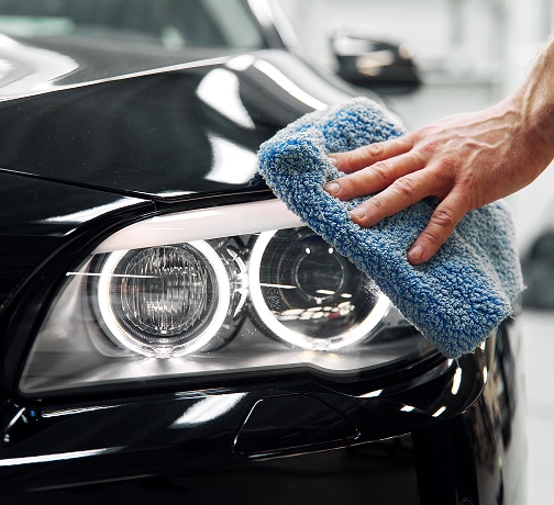 Lustrage de Phare de Voiture : Entretien et Brillance Main polissant un phare de voiture noir brillant avec un chiffon microfibre bleu texturé pour un entretien automobile.
