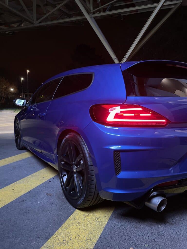 Scirocco R Bleu Nuit : Échappement Akrapovic Scirocco R bleu vif garé de nuit. Arrière sportif, feux LED allumés et embout d'échappement Akrapovic. Jantes noires.