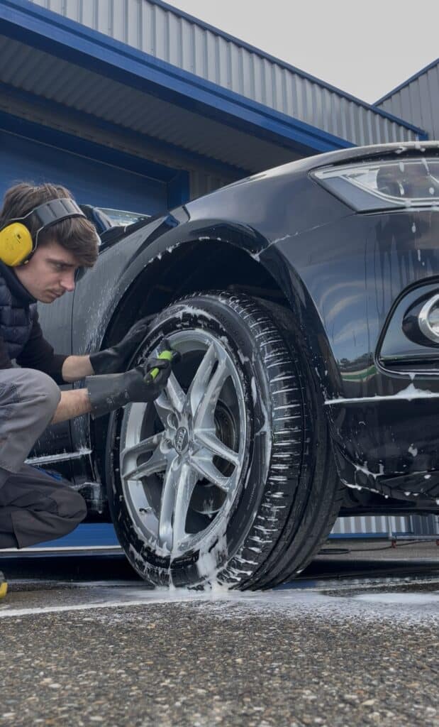 Détailing auto : nettoyage professionnel des jantes Audi. Detailer en protection auditive nettoyant une jante Audi noire couverte de mousse avec une brosse. Lavage auto manuel et précis.