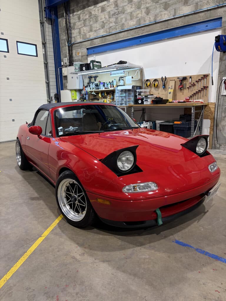 Mazda Miata NA rouge : l'icône des phares escamotables. Superbe Mazda Miata NA rouge vif, phares escamotables levés, garée dans un atelier. Jantes chromées et étagères d'outils.